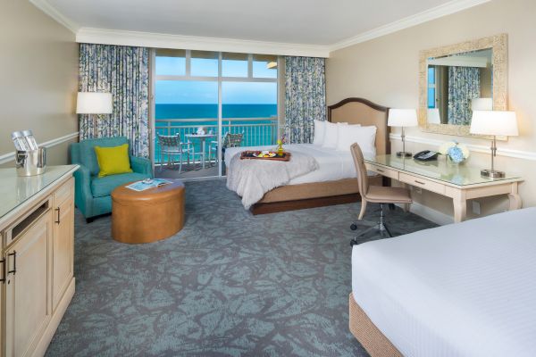 The image shows a hotel room with two beds, a desk, a mirror, and a seating area. There is a balcony with an ocean view.
