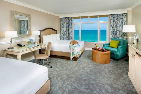 The image shows a hotel room with two beds, a desk, a chair, a comfy seat, and large windows with an ocean view, decorated in a coastal theme.