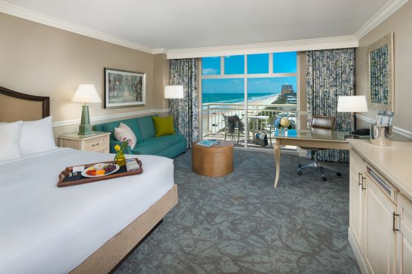 A hotel room features a bed, sofa, desk, and a balcony with an ocean view. There's a tray of food on the bed and a vibrant interior.