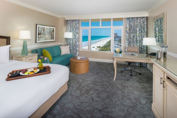 A bright hotel room with an ocean view, featuring a bed, seating area, desk, and tray with fruit. The window overlooks a beach and ocean.