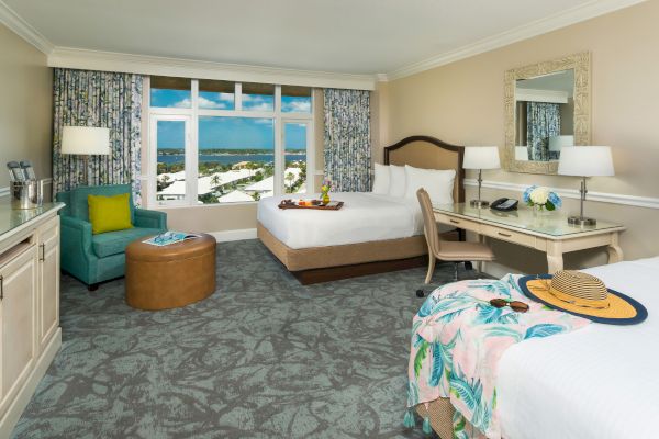 A hotel room with two beds, a desk, a teal chair, a window with a scenic view, and a light, bright decor.