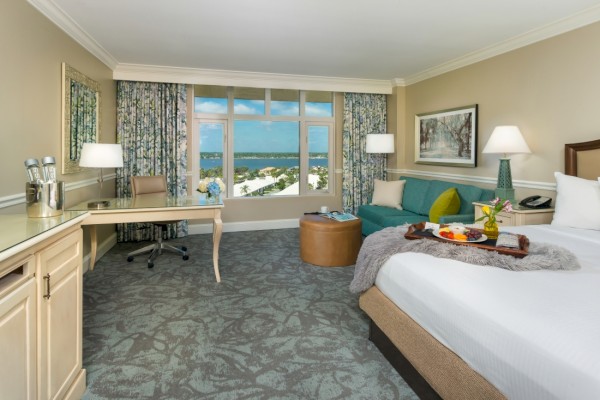 This is a hotel room with a large window showing a scenic view of water and greenery, a bed with breakfast, a sitting area, and a desk.