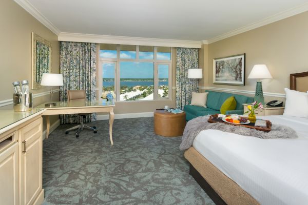 A hotel room featuring a bed, desk, sofa, and a window with an ocean view; a tray with food on the bed ends the sentence.