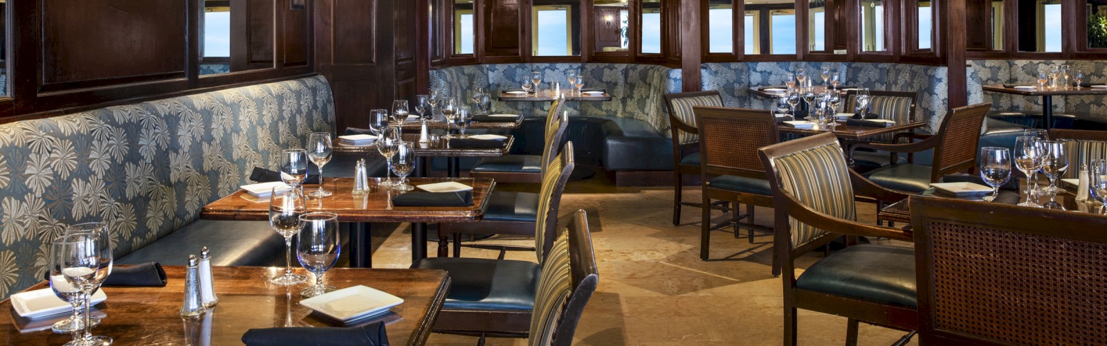 An upscale restaurant with wooden furniture, upholstered seats, neatly set tables, and a ceiling fan under a wooden-beamed ceiling.