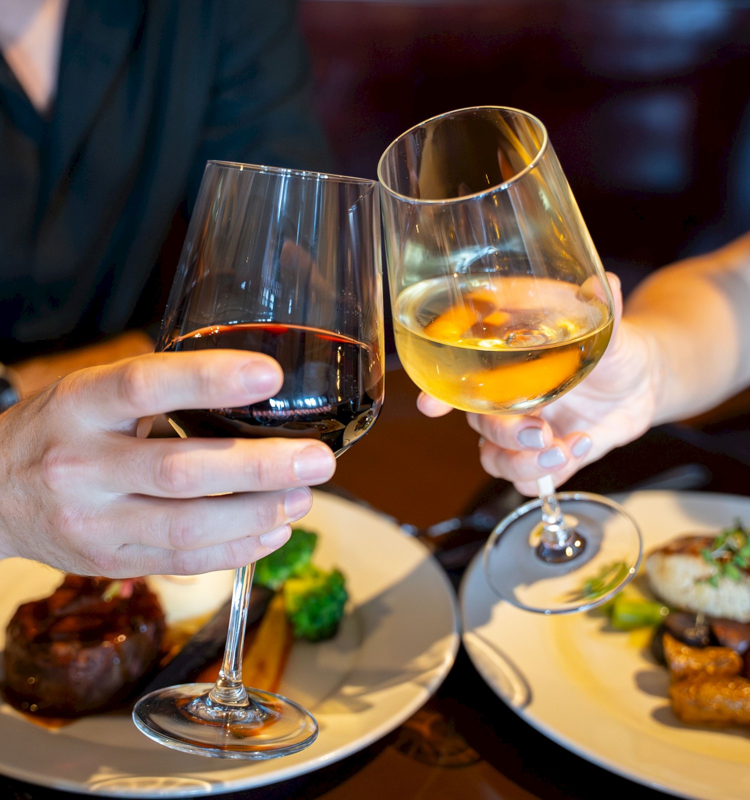 Two people clink wine glasses over plated meals in a cozy dining setting, sharing a toast and enjoying a meal together.