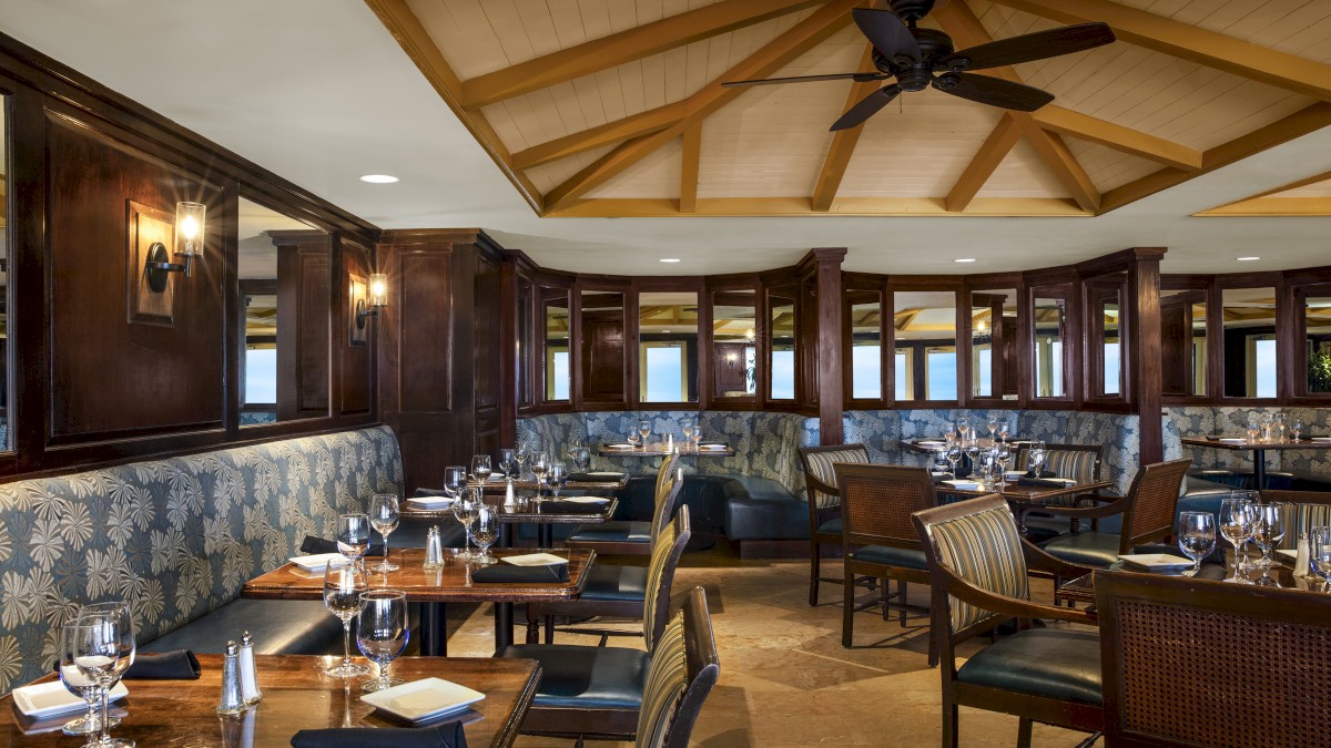 A warmly-lit restaurant with wooden furniture, cushioned seating, and set tables with glassware and napkins, under a ceiling fan and wooden beams.