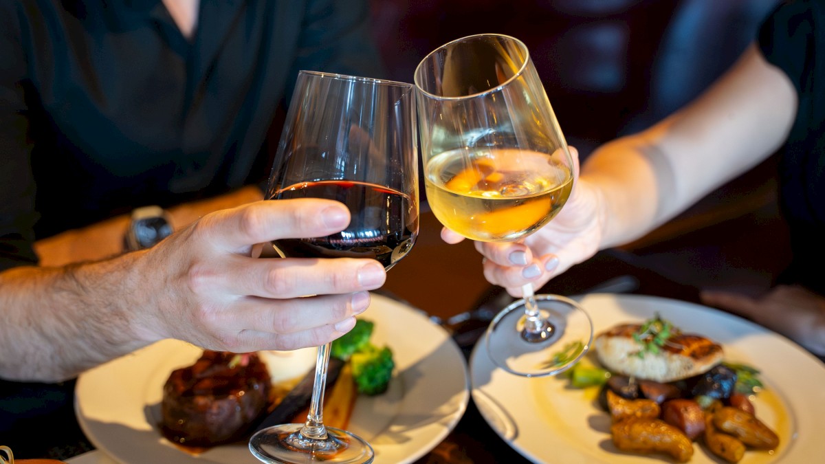 Two people clink glasses of red and white wine over a dinner table with dishes of meat, potatoes, and vegetables.