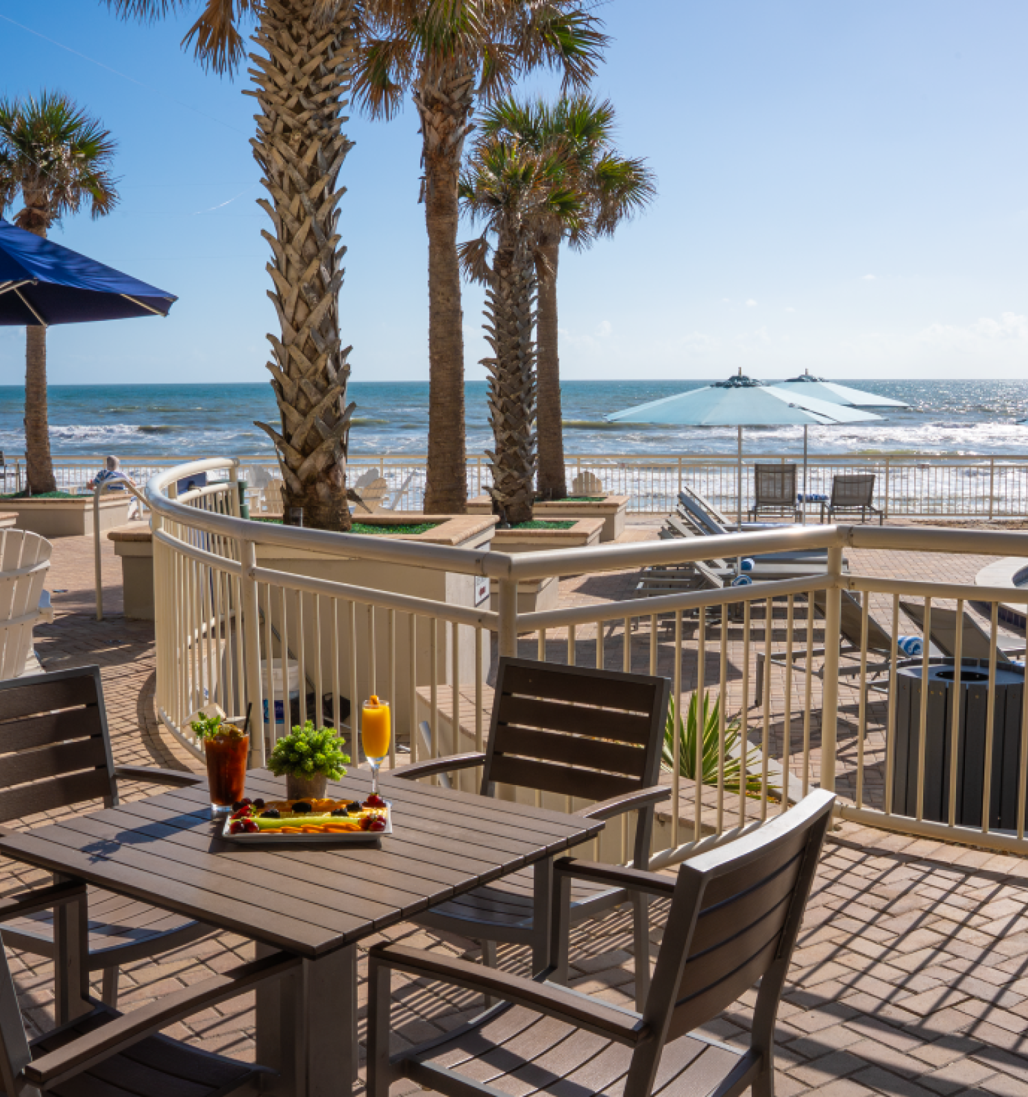 A beachside patio with tables, chairs, umbrellas, palm trees, and a view of the ocean and a small pool.