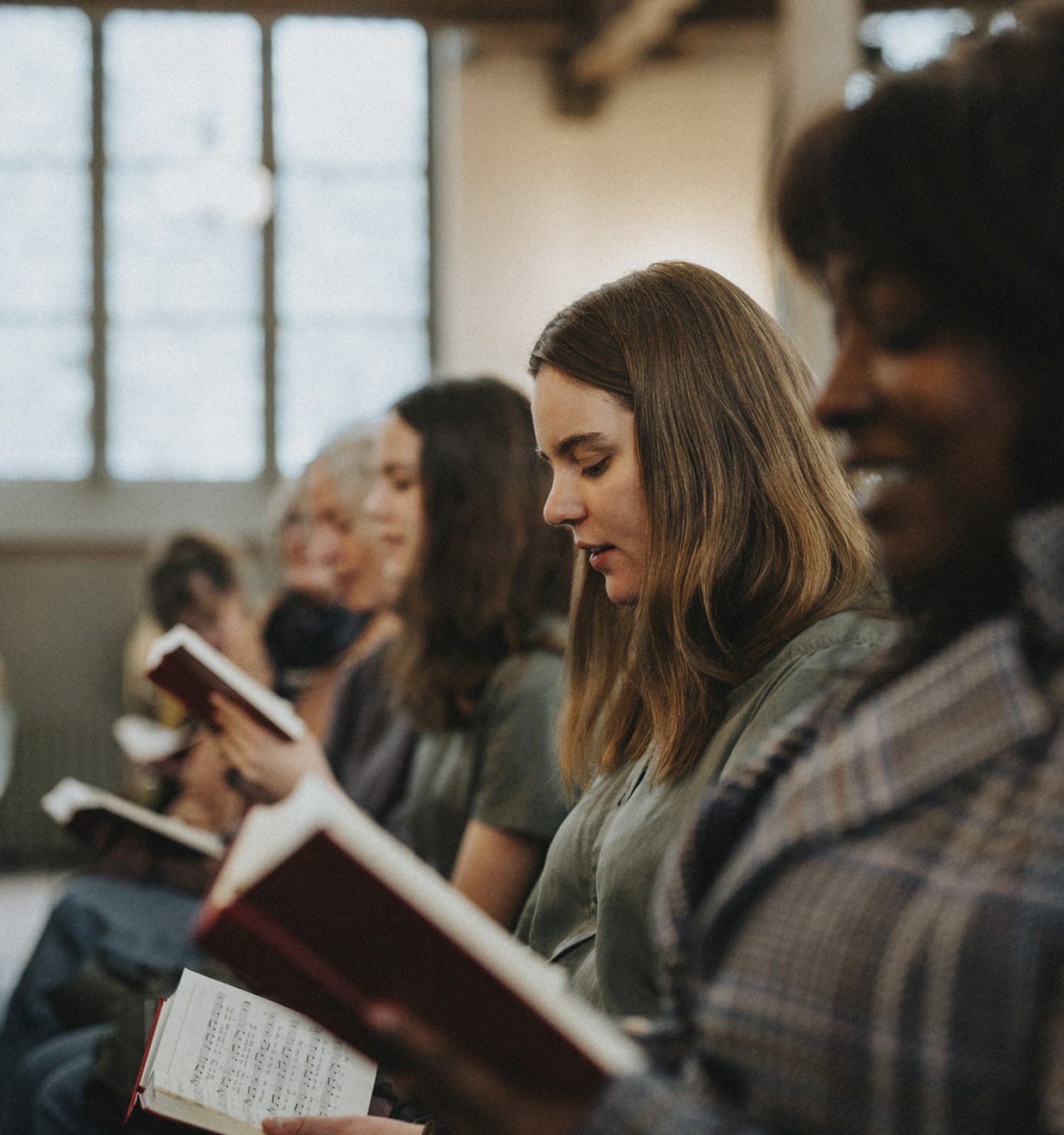 A diverse group of people seated in a church or classroom, reading books or hymnals, focused on their pages amidst a bright, airy room.