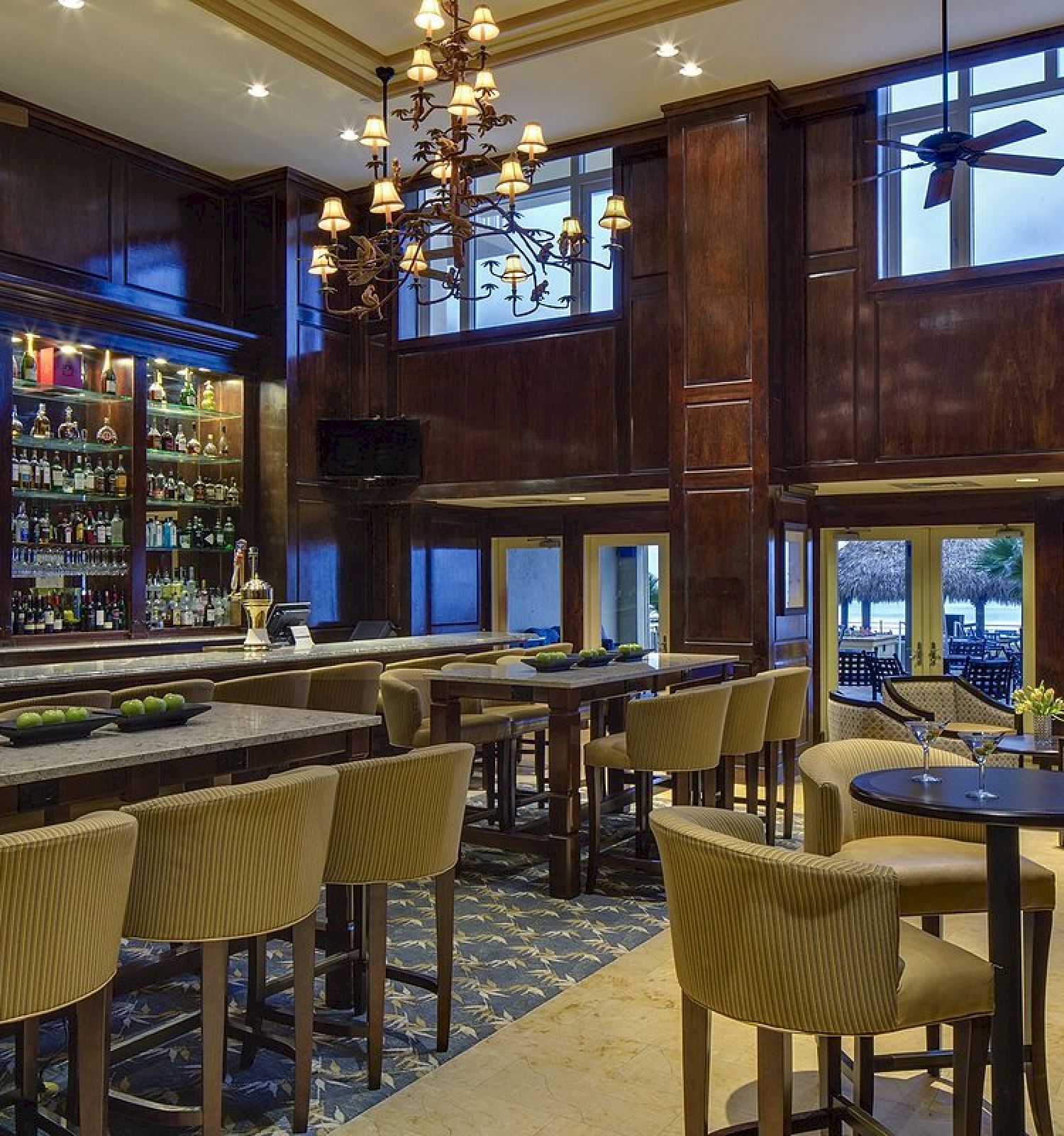 The image depicts a well-furnished bar with high stools, tables, a chandelier, a stocked bar, and large windows looking out to a scenic view.