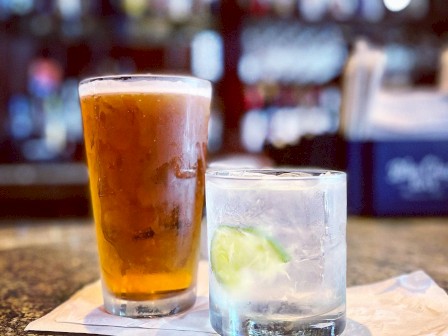 Two drinks on a bar counter: a tall glass of iced tea or beer and a glass with a lime, on a napkin, blurred bar background.