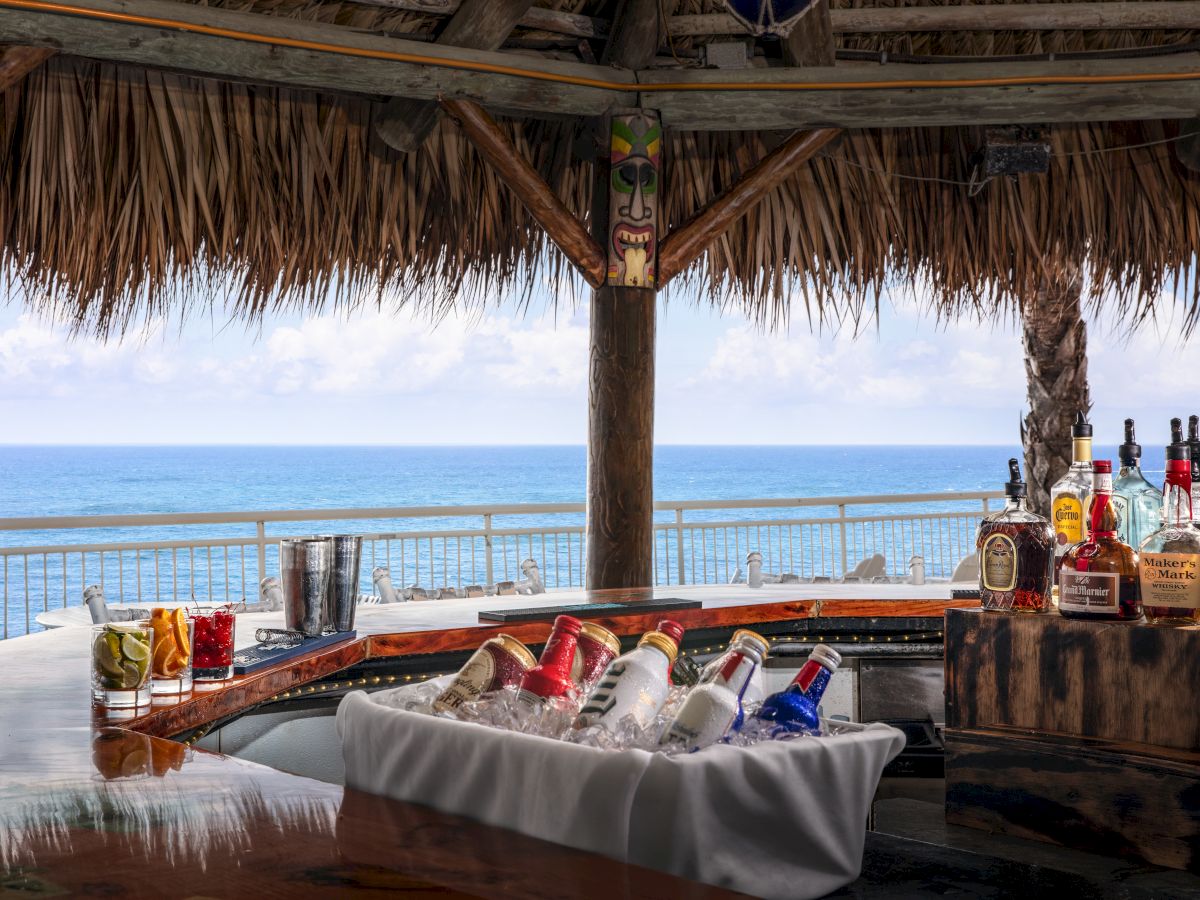 A tropical beach bar with various alcoholic beverages displayed, and an ocean view in the background.