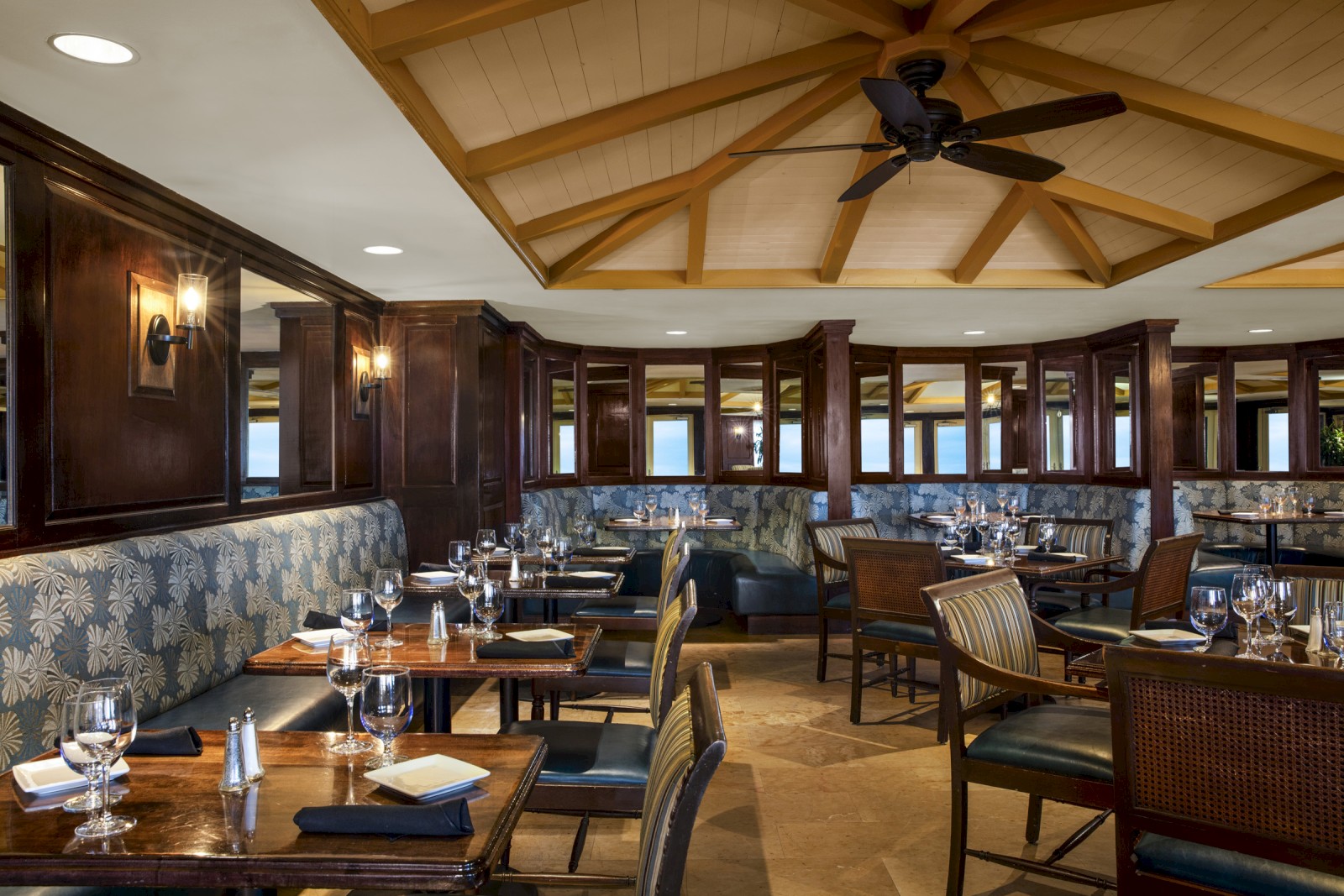 The image shows an elegant restaurant interior with wooden furnishings, cushioned seating, neatly arranged tables, and a ceiling fan.