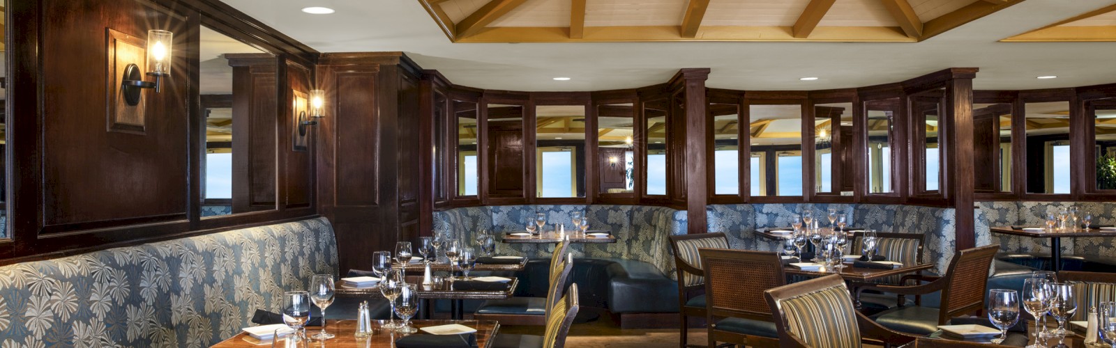 The image shows an elegant restaurant interior with wooden furnishings, cushioned seating, neatly arranged tables, and a ceiling fan.