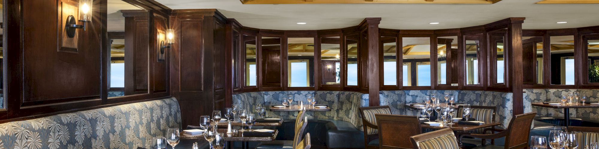 The image shows an elegant restaurant interior with wooden furnishings, cushioned seating, neatly arranged tables, and a ceiling fan.