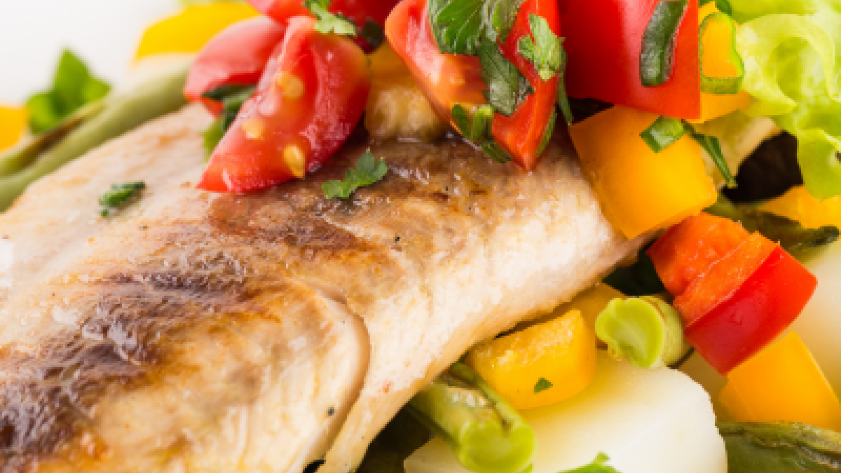 The image shows a plate with a piece of grilled fish, garnished with chopped tomatoes, parsley, and surrounded by various vegetables, including bell peppers.