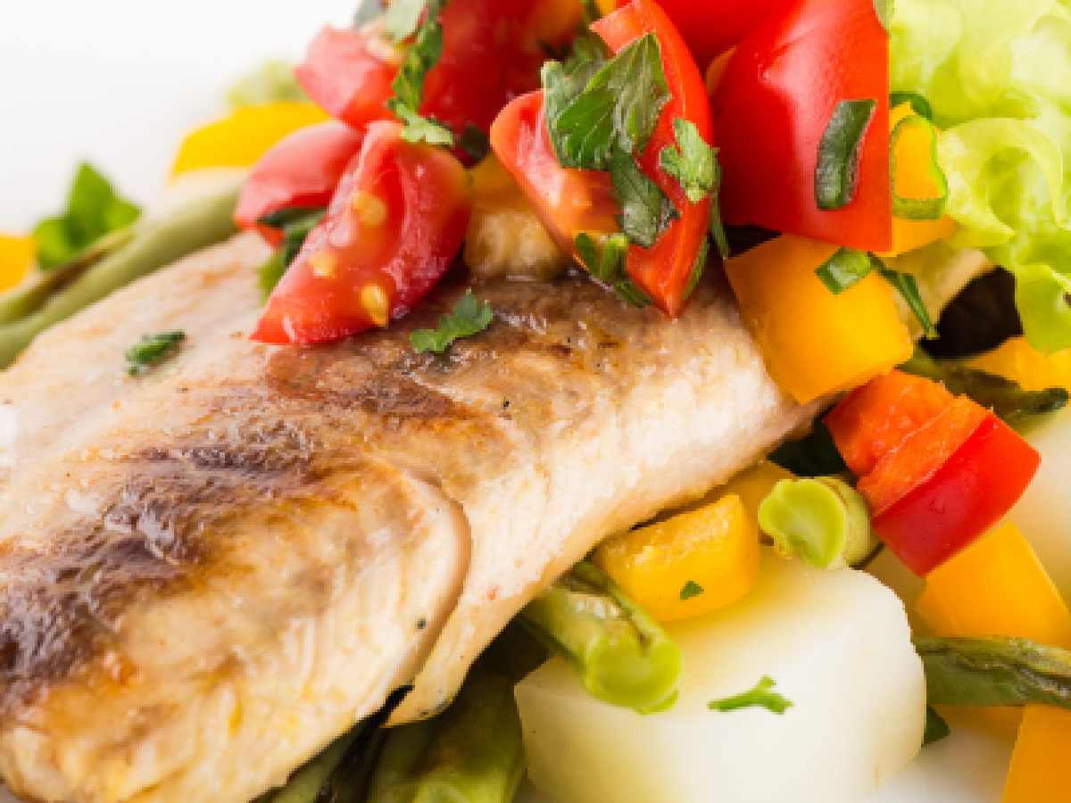 The image shows a plate with a piece of grilled fish, garnished with chopped tomatoes, parsley, and surrounded by various vegetables, including bell peppers.