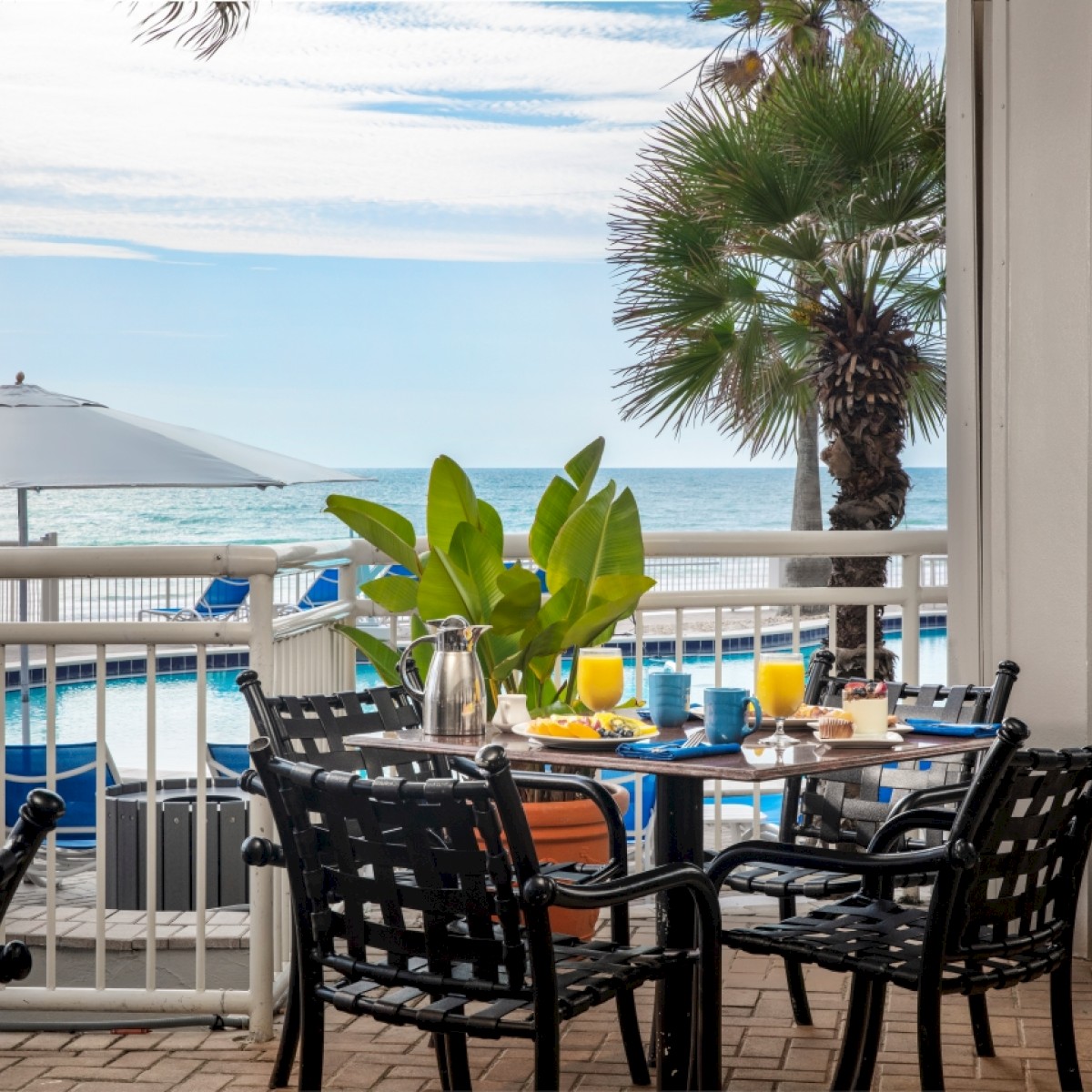 A breakfast setup on a patio overlooking a pool, palm trees, and the ocean with blue sky and scattered clouds.