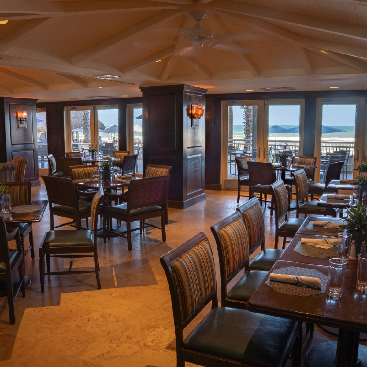 A cozy seaside restaurant with wooden tables, colorful chairs, and a view of the beach and ocean outside.