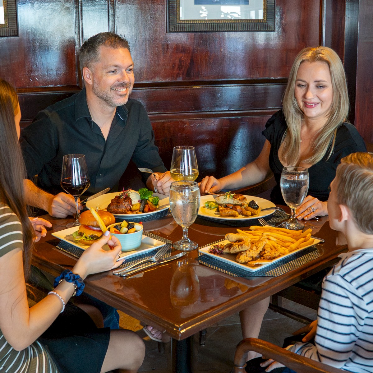 A family of four enjoys a meal at a restaurant, with wine, water, and dishes of various foods on the table.