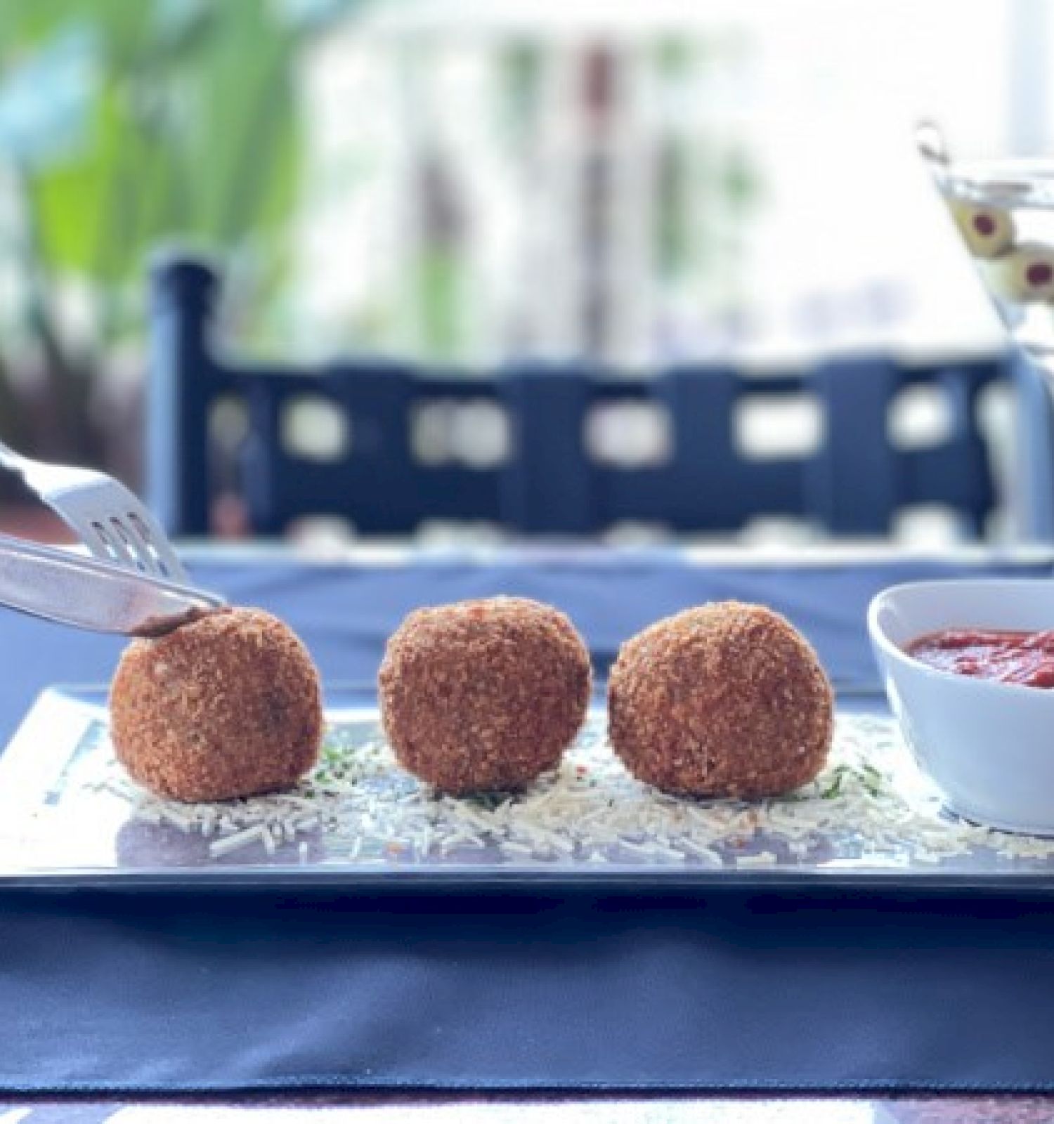 Three breaded balls on a plate with a dipping sauce and a drink garnished with olives. A hand is cutting one of the balls.