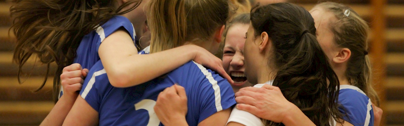 A group of girls in blue jerseys huddle in a circle, arms around each other, celebrating with smiles after a game.