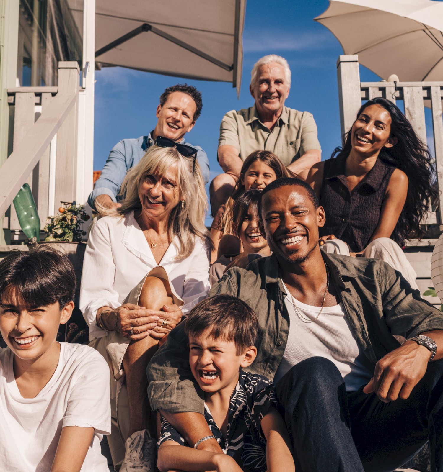 A big, smiling multi-generational family group posing on a sunny outdoor staircase, with pastel buildings and umbrellas in the background.