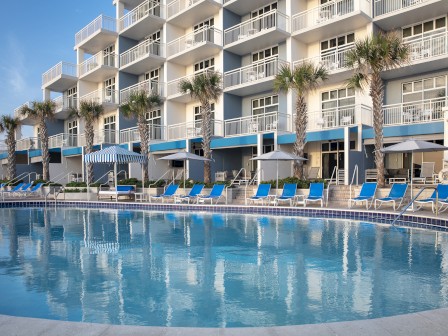 A sunny hotel pool area with blue lounge chairs, palm trees, and white balconies overlooking the water, perfect for a vacation.