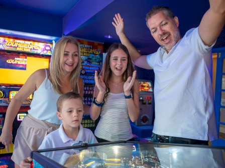 A family celebrates at an arcade, smiling near a pinball table while a boy sits at a game, and the parents cheer in a colorful arcade.