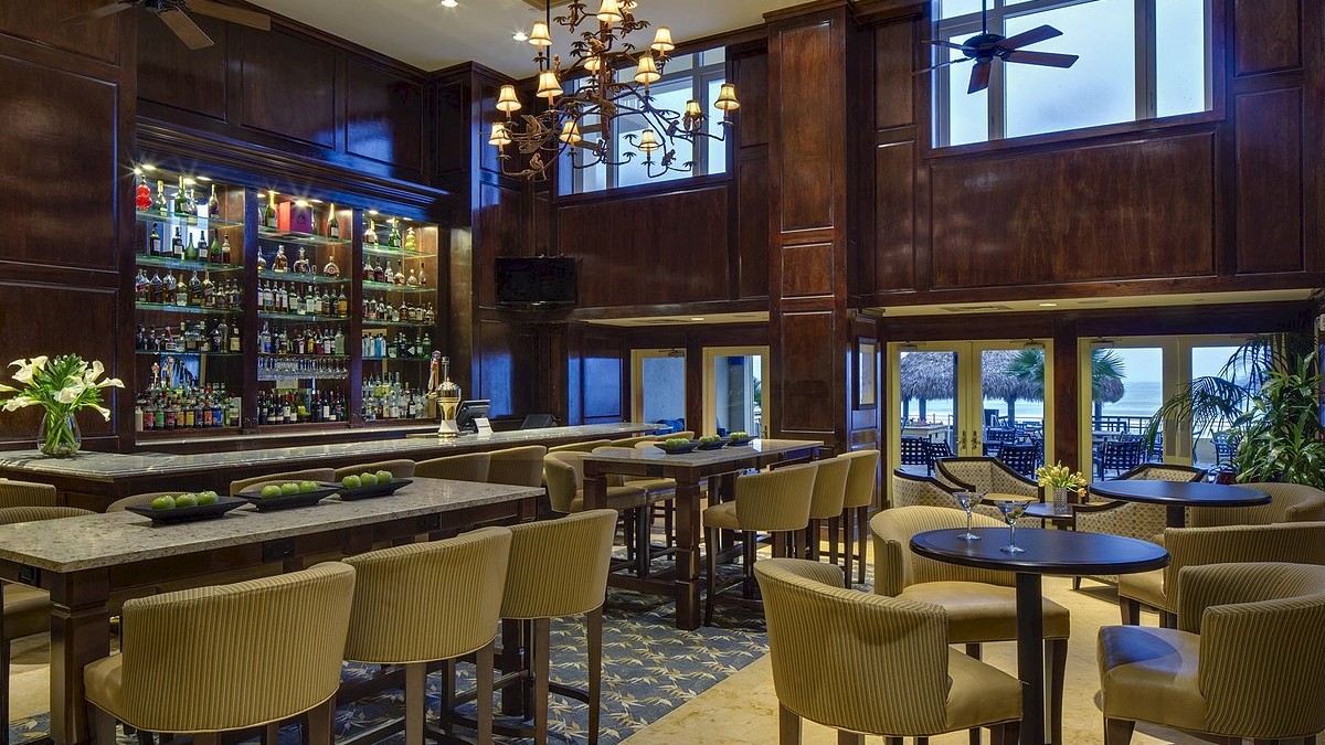 A modern bar with beige chairs, tables, a chandelier, a well-stocked bar, and large windows overlooking a scenic view.