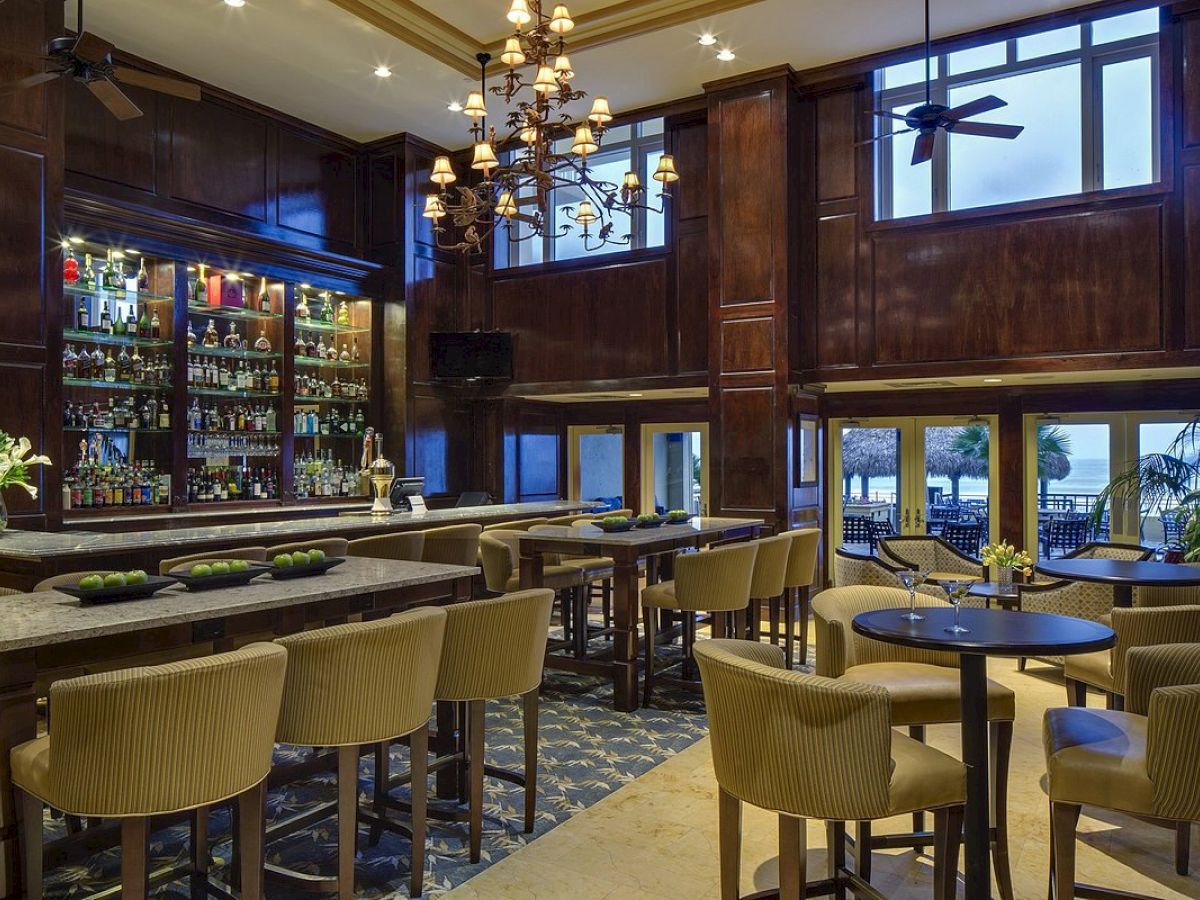 A modern bar with beige chairs, tables, a chandelier, a well-stocked bar, and large windows overlooking a scenic view.