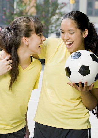 A group of girls in yellow shirts chats and laughs outdoors, one girl holds a soccer ball while another carries a green backpack.