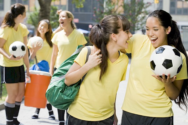 A group of girls in yellow shirts chats and laughs outdoors, one girl holds a soccer ball while another carries a green backpack.