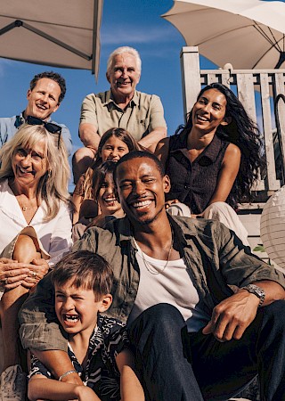 A diverse, smiling group of family and friends posing on a sunny outdoor porch, summer vibe with chairs, plants, and umbrellas.