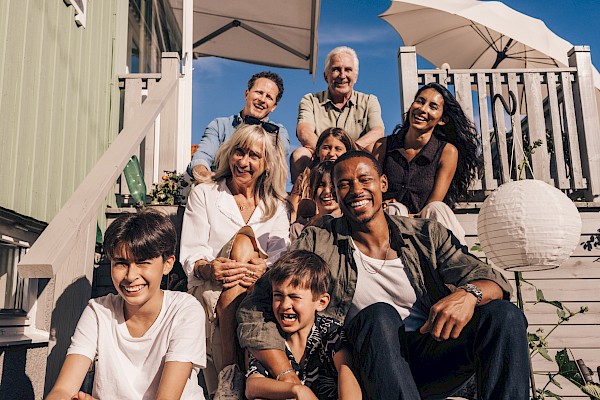 A diverse, smiling group of family and friends posing on a sunny outdoor porch, summer vibe with chairs, plants, and umbrellas.