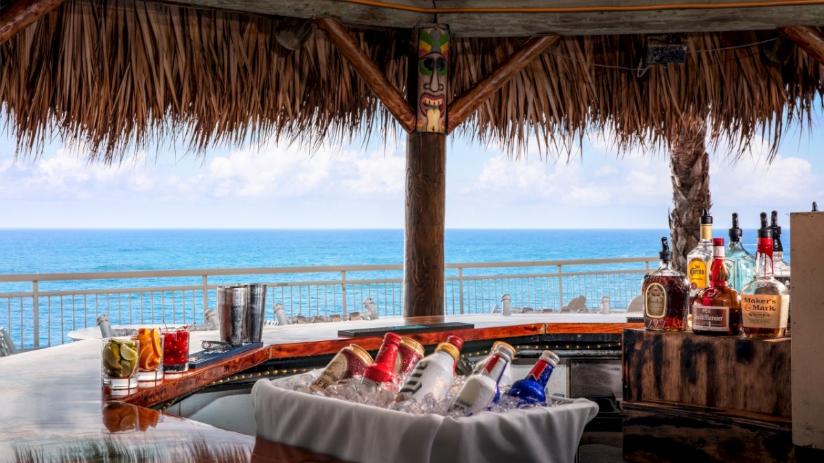 A tropical beach bar with alcohol bottles, drinks on the counter, overlooking the ocean under a thatched roof.
