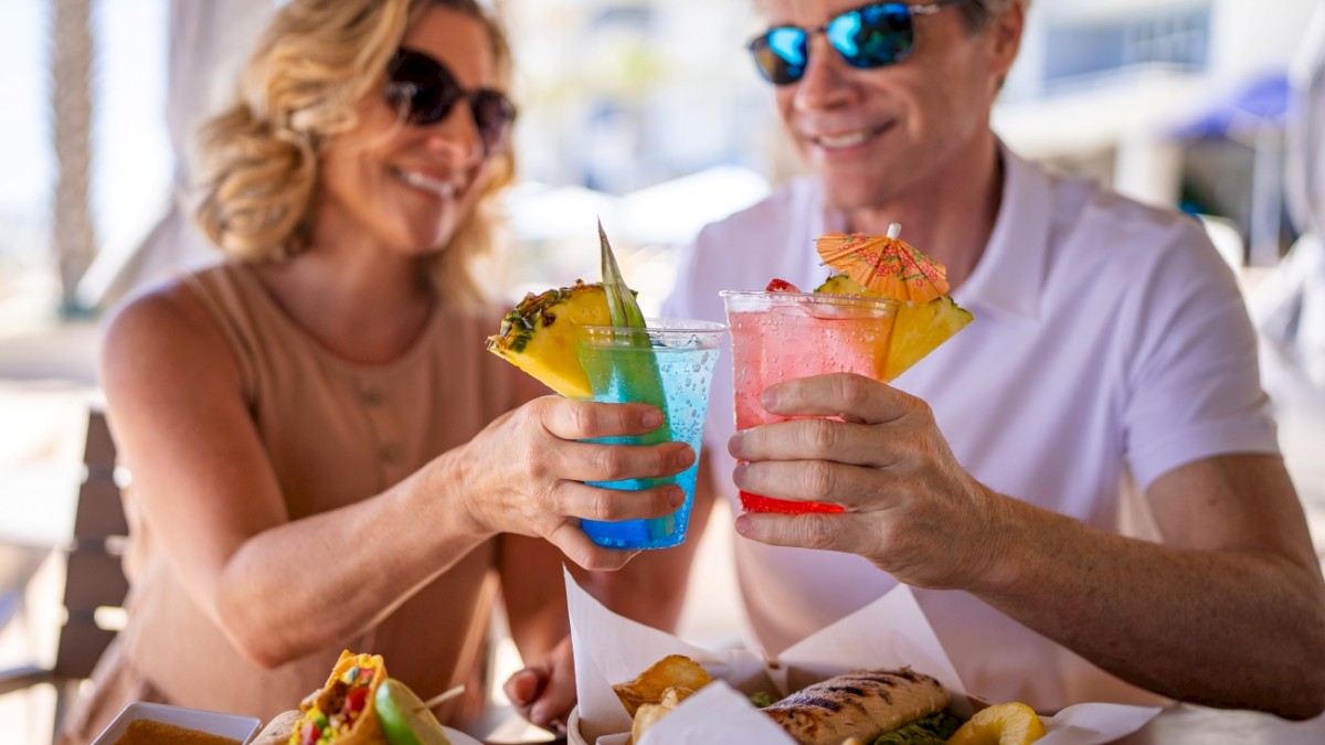A smiling couple wearing sunglasses toasting colorful tropical drinks with pineapple garnishes at a sunny outdoor restaurant.