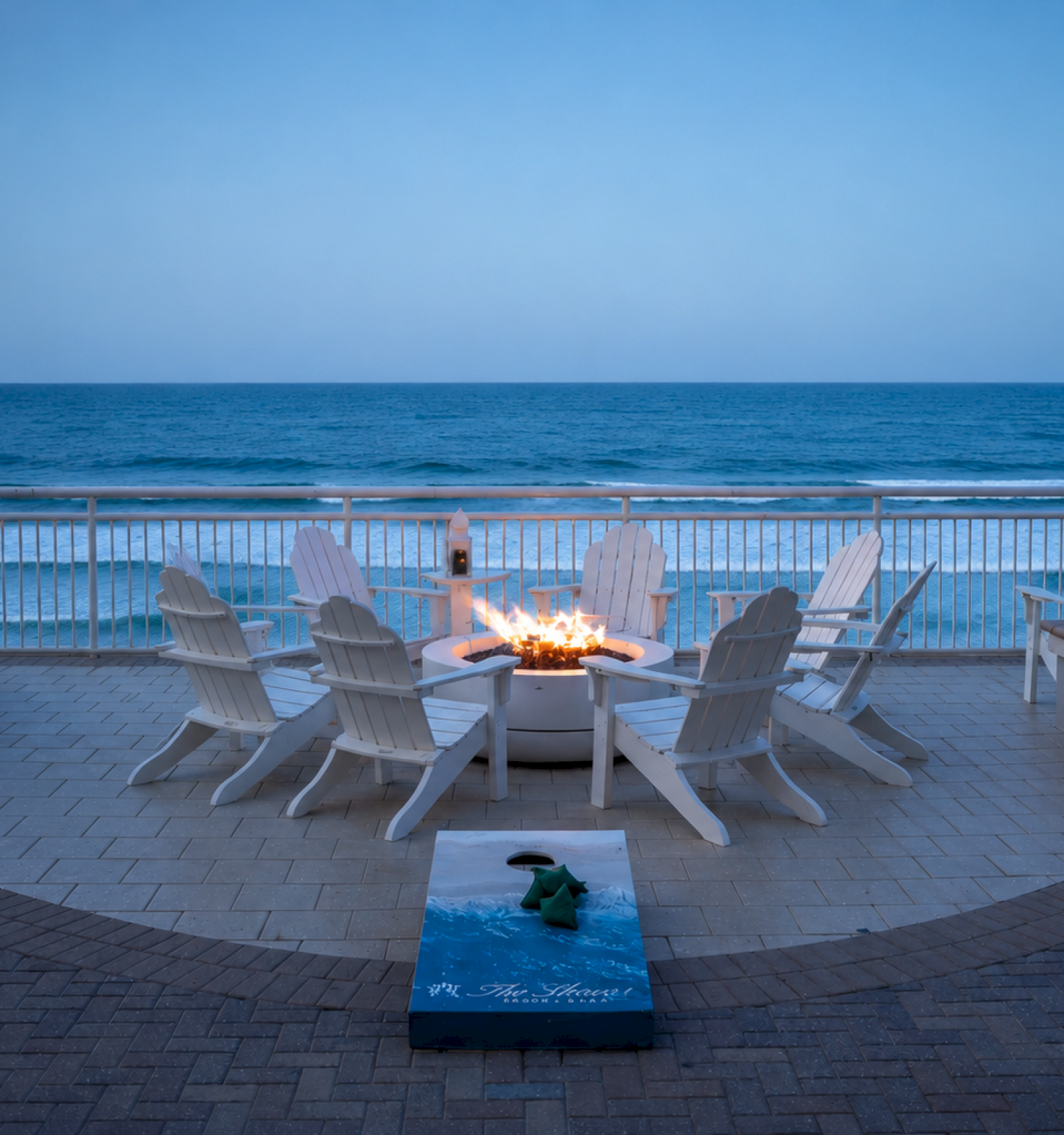 A serene oceanfront scene with white chairs around a firepit on a patio, overlooking the ocean at dusk, creating a cozy, relaxing atmosphere.