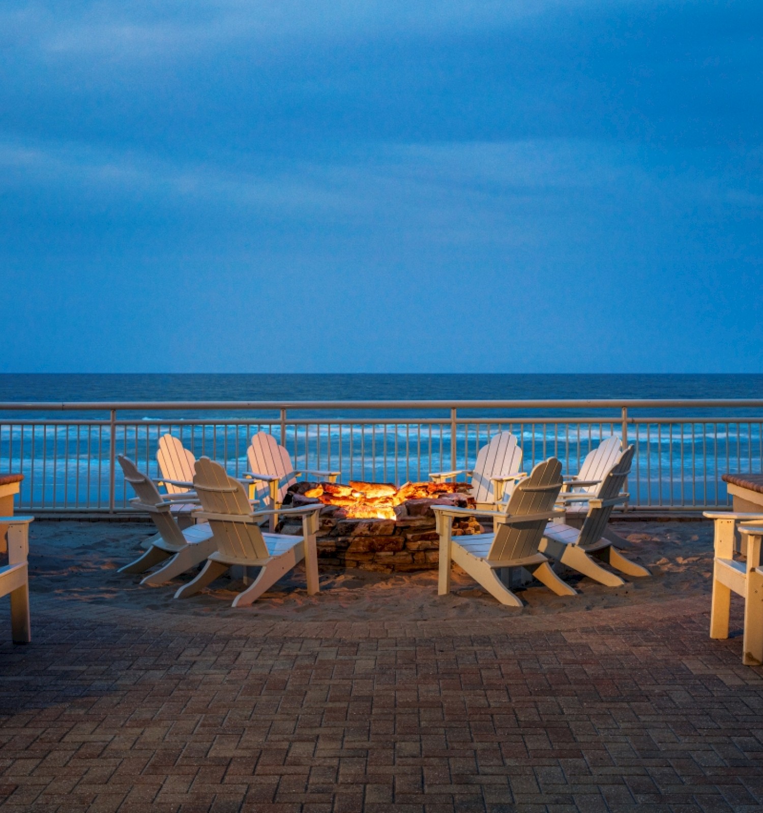 Beach scene with white chairs around a fire pit, palm trees, and ocean view against a blue sky.