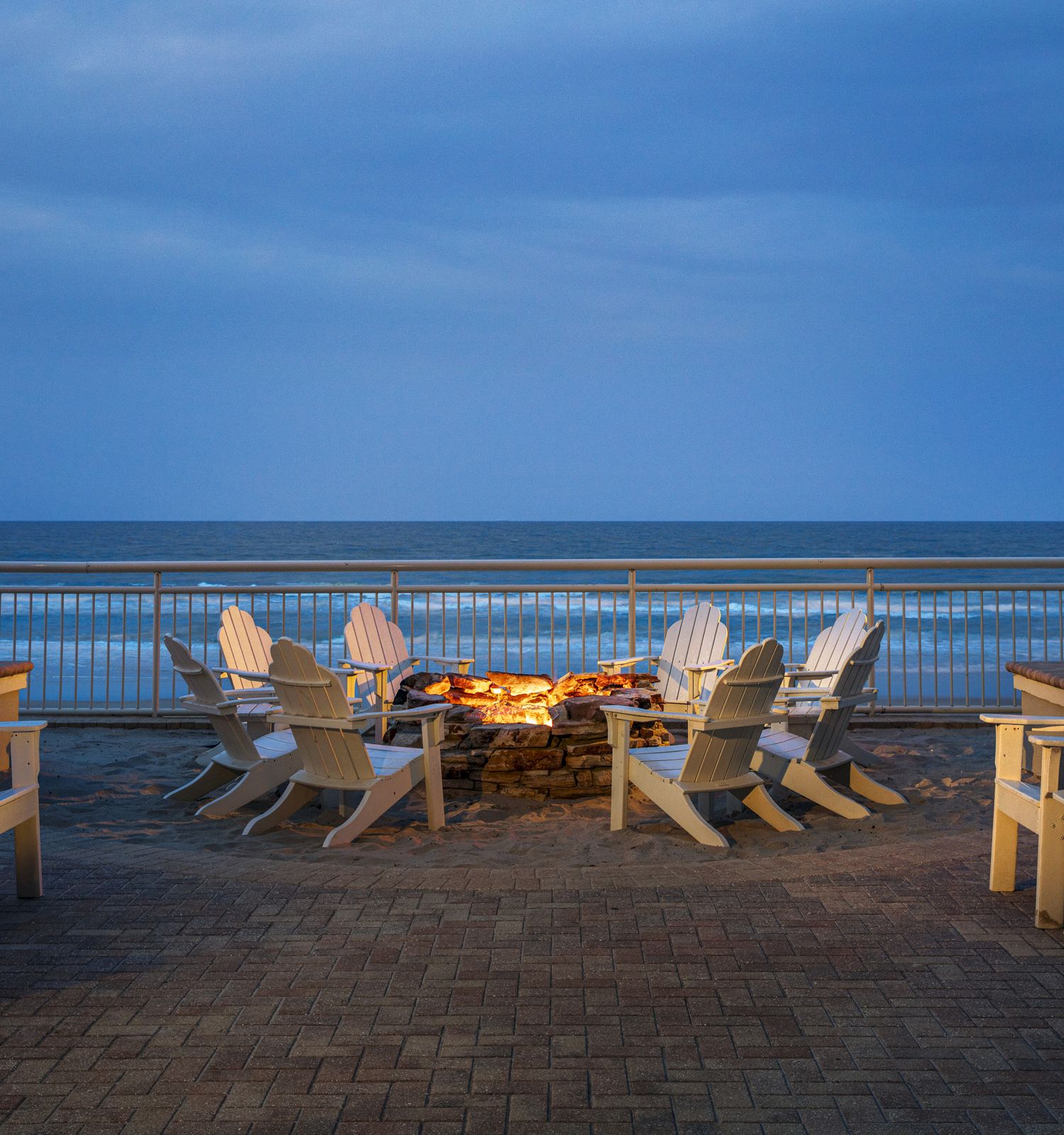 The image shows a cozy beach setting with white Adirondack chairs arranged around a lit fire pit, flanked by two palm trees, overlooking the ocean.