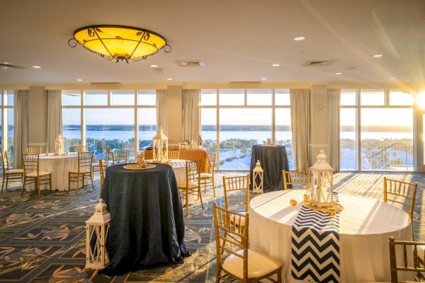 Elegant banquet room with round tables, lantern centerpieces, and large windows showing a river view at sunset.