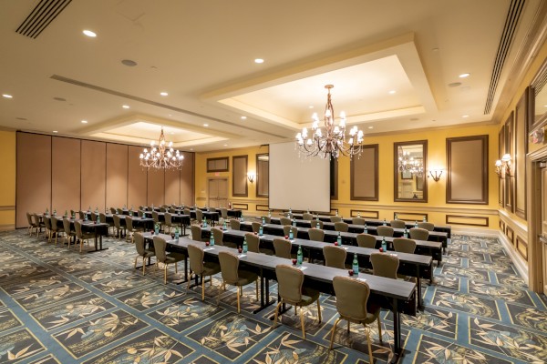 A spacious conference room set for a seminar with long rows of tables and chairs, chandeliers, ornate walls, and patterned carpet, ready for attendees.