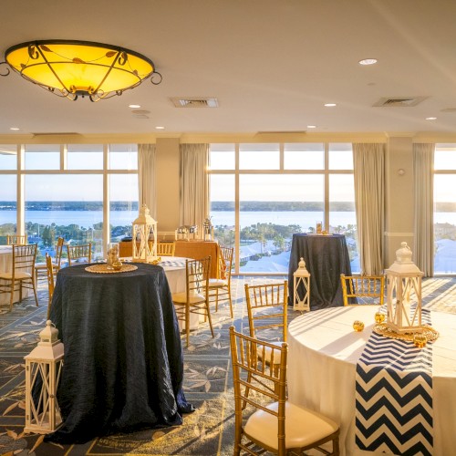 A sunlit banquet room with round tables, white linens, decorative lanterns, and a sea view through large windows.