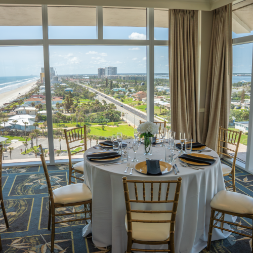 Elegant table set for a meal with a view of a sunny beach and green cityscape visible through large windows.