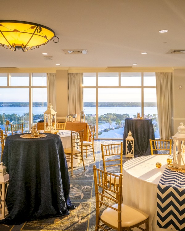 A sunlit banquet room with round tables, white linens, decorative lanterns, and a sea view through large windows.