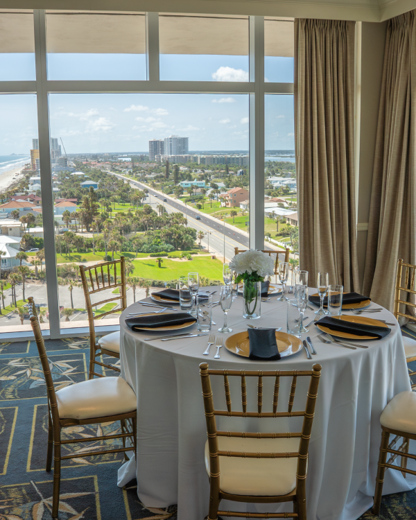 Elegant table set for a meal with a view of a sunny beach and green cityscape visible through large windows.