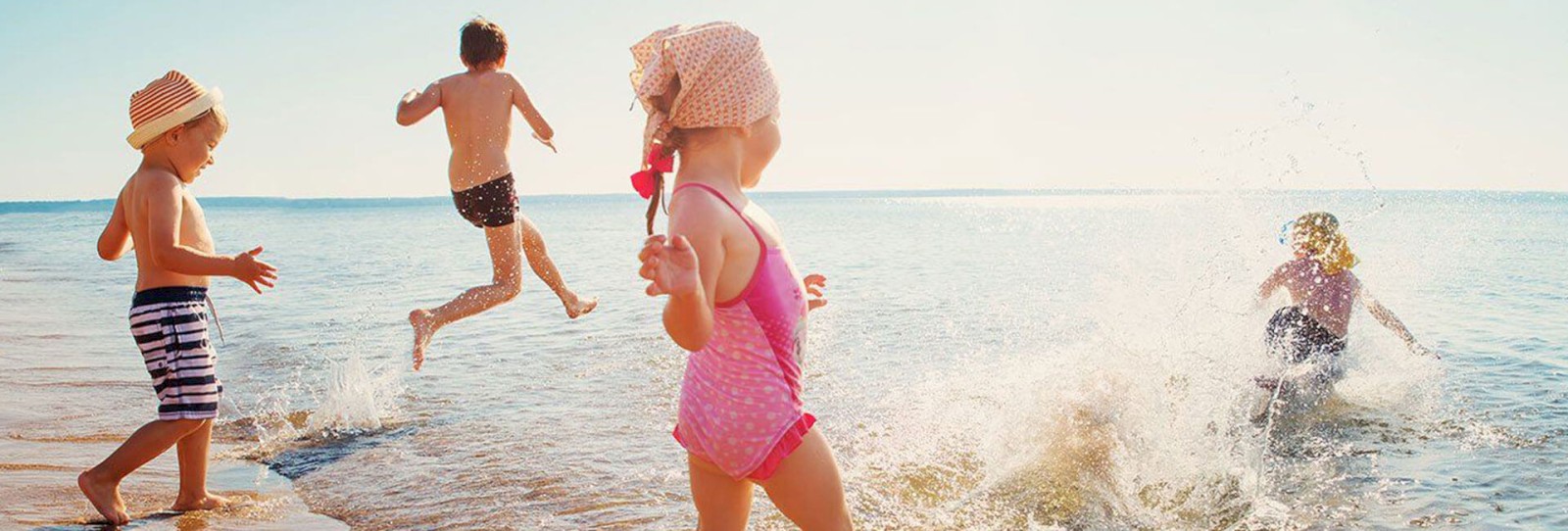 Four children are playing at the beach, splashing water and enjoying the sun, with one child jumping into the water, creating a splash.
