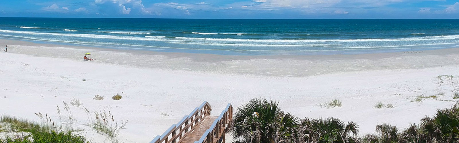 A serene beach scene with white sand, blue ocean waves, and a wooden walkway leading to the beach. Only a few people are visible.