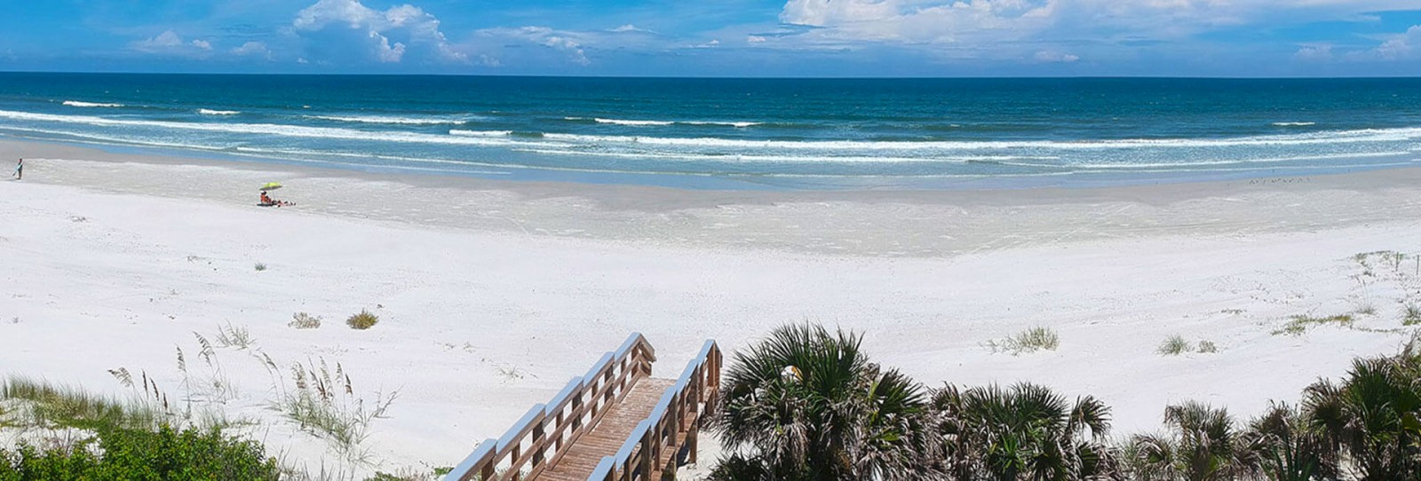 A serene beach scene with white sand, blue ocean waves, and a wooden walkway leading to the beach. Only a few people are visible.