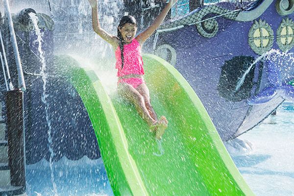 A child in a pink swimsuit is sliding down a green water slide with arms raised and water splashing around, likely in a water park.